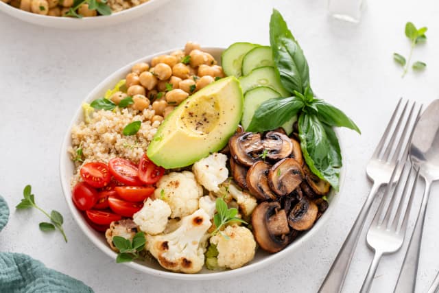 A vegetarian lunch bowl with mushrooms, baby tomatos, cauliflowwer, avacado, cucumber, chic peas, couscous, and more